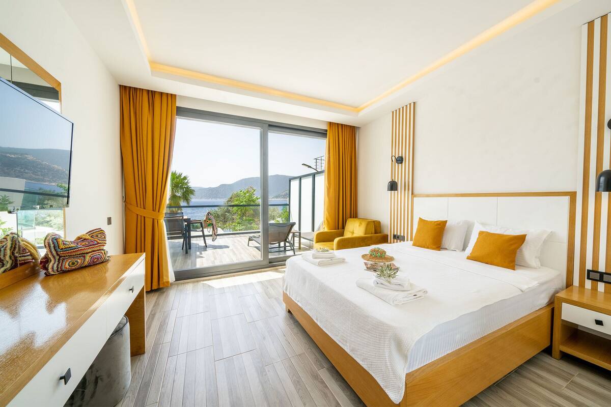 A modern hotel room with a large bed, a flat-screen TV, a balcony with yellow curtains, and a view of the mountains.