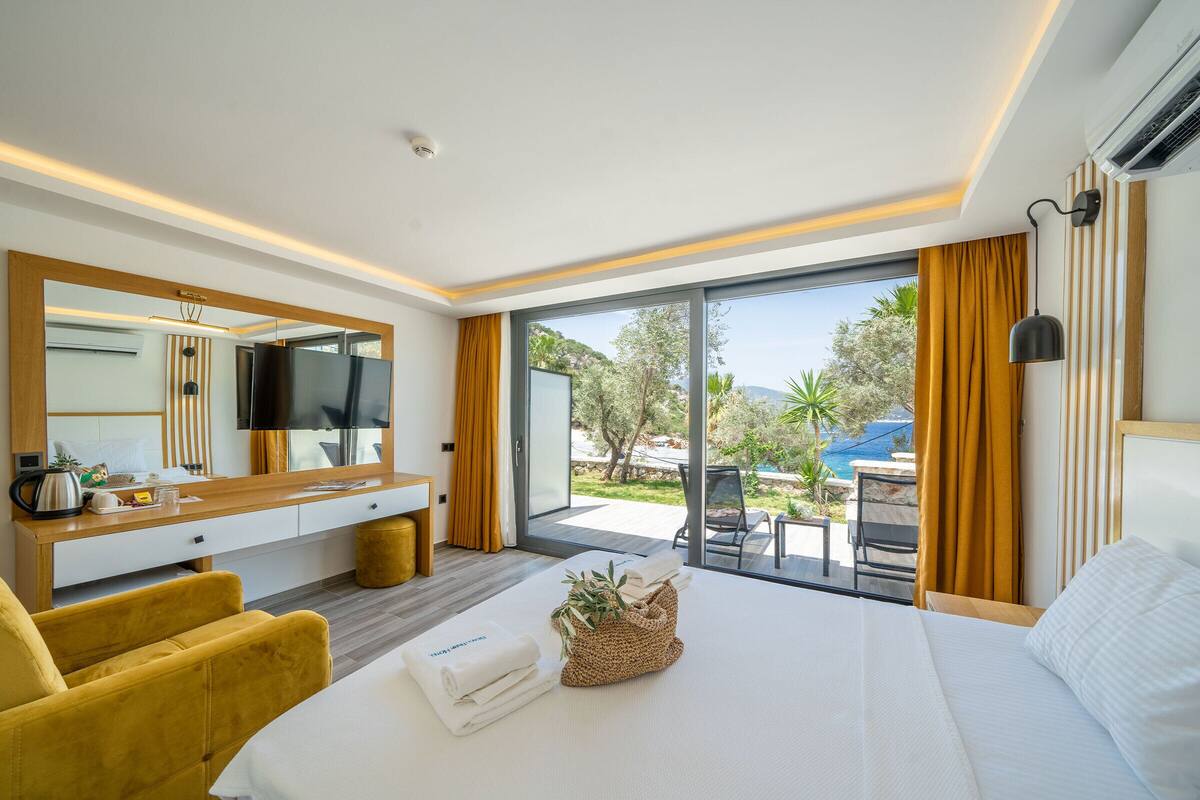 A hotel room with a bed, a yellow armchair, a large mirror, a flat-screen TV, a wooden desk, and a view of the outdoors.