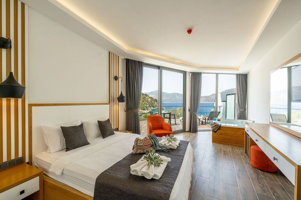 A modern hotel room with a large bed, a desk, a chair, and a view of the sea.