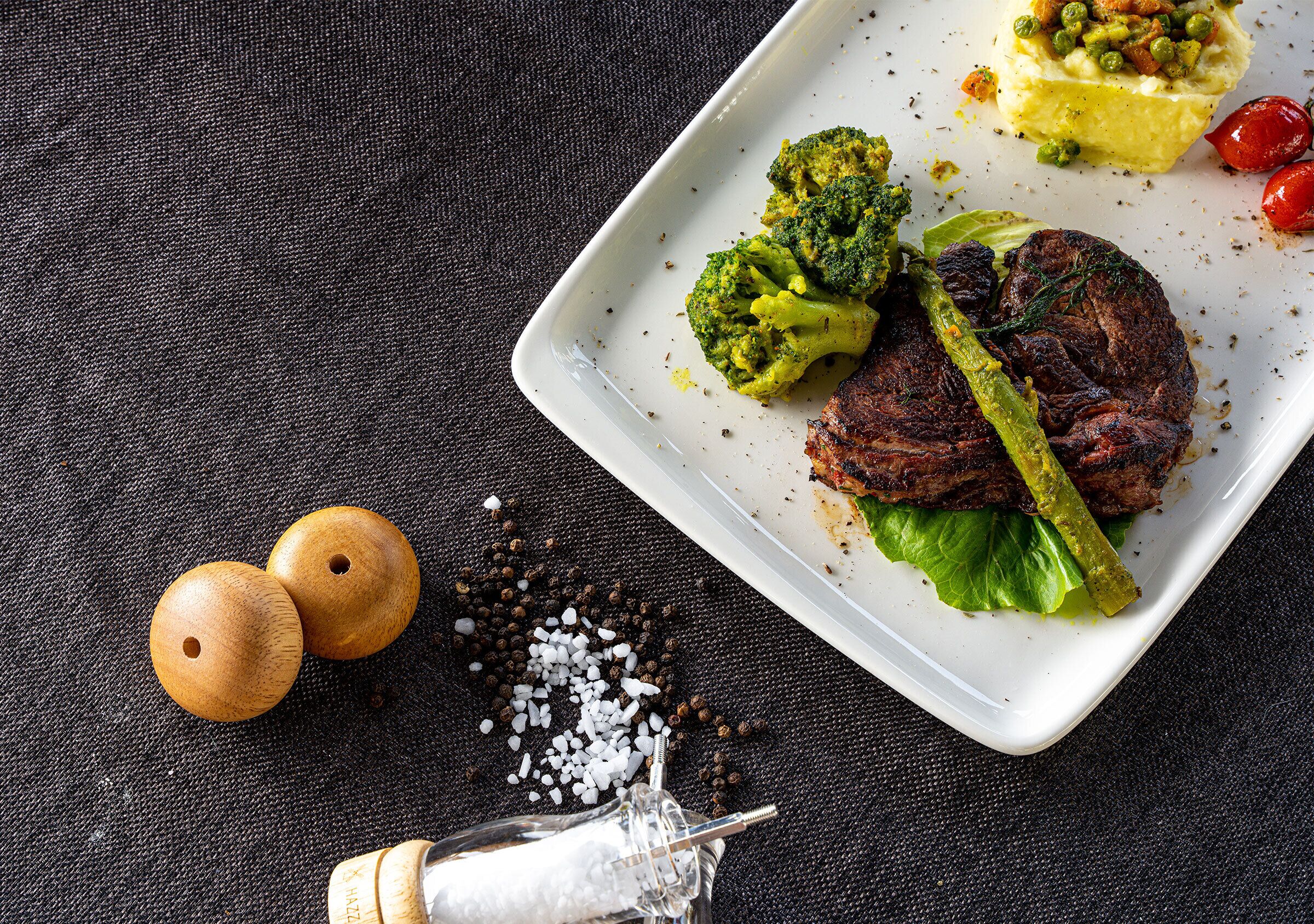 A white plate with a steak, broccoli, and a green sauce, set on a dark textured tablecloth.