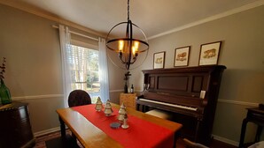 A dining room with a wooden table, red tablecloth, and three glass Christmas trees. A piano is against the wall, and a chandelier hangs above the table.