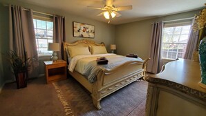 A bedroom with a large bed, a ceiling fan, a nightstand with a lamp, a dresser, and a window with curtains.