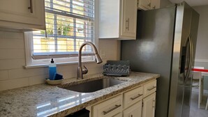 A kitchen with white cabinets, a stainless steel refrigerator, a granite countertop, and a window with blinds.