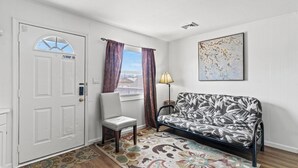 A room with a white door, a patterned sofa, a chair, a lamp, and a framed artwork on the wall.