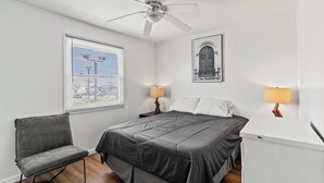 A bedroom with a bed, a chair, a ceiling fan, a window with blinds, and a wall-mounted lamp.
