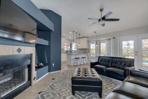 Cozy living space with a fireplace, leather sofa, and dining area, featuring a ceiling fan and large windows.