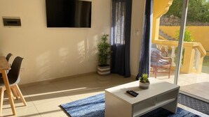 Ein Wohnzimmer mit Fernseher, Sofa, Couchtisch, einer Pflanze und einem Balkon mit Stuhl.