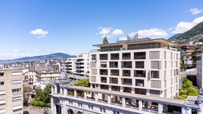 A modern multi-story building with balconies and large windows, surrounded by other buildings and a mountainous backdrop.