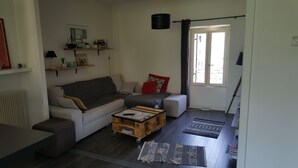 Een woonkamer met een bank, een salontafel en een boekenkast. Er is een raam met gordijnen en een deur.