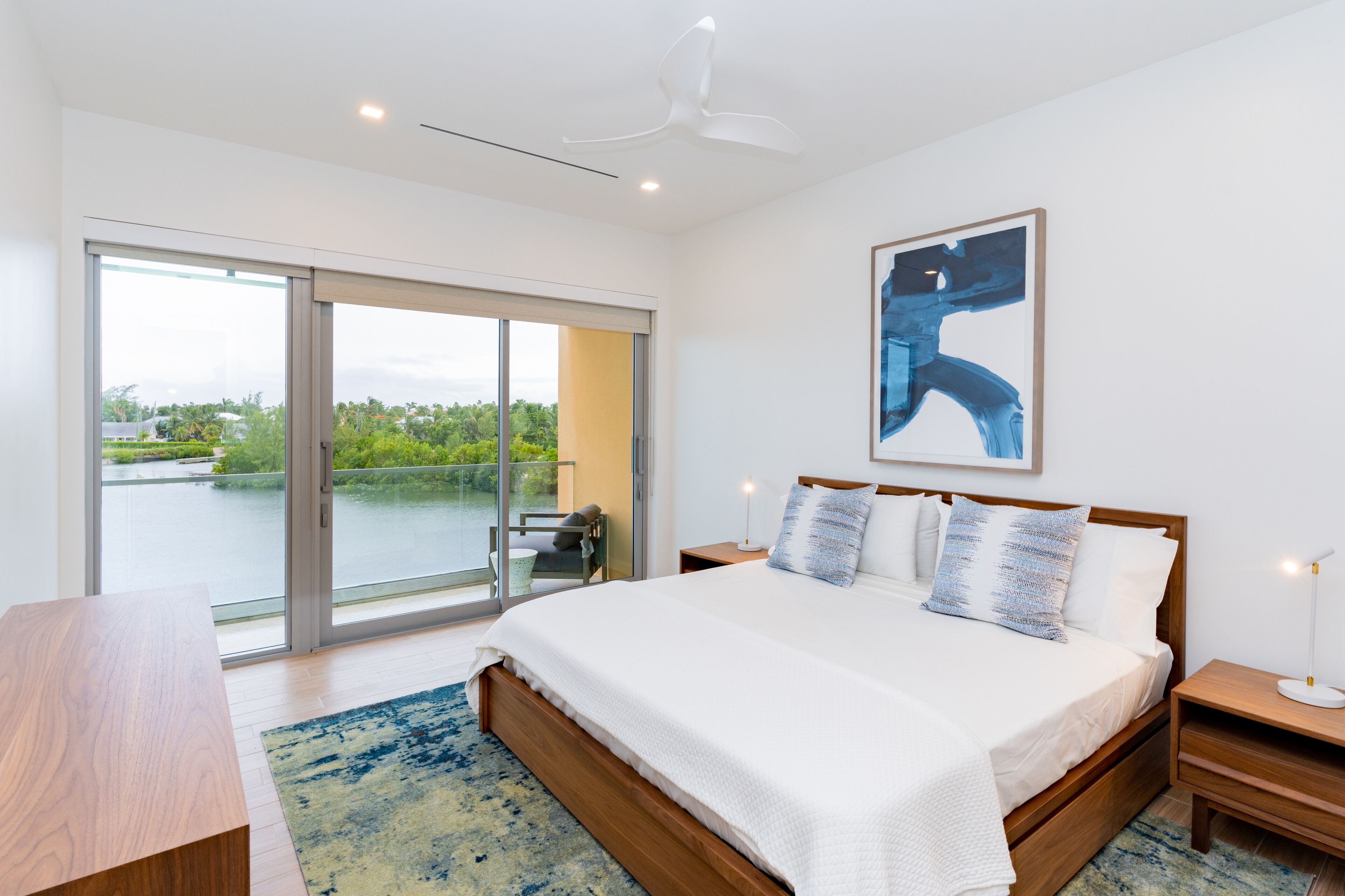 A modern bedroom with a large bed, a wooden nightstand, a desk, and a view of a balcony with a glass door.