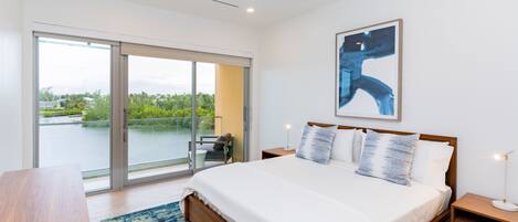 A modern bedroom with a large bed, a wooden nightstand, a desk, and a view of a balcony with a glass door.