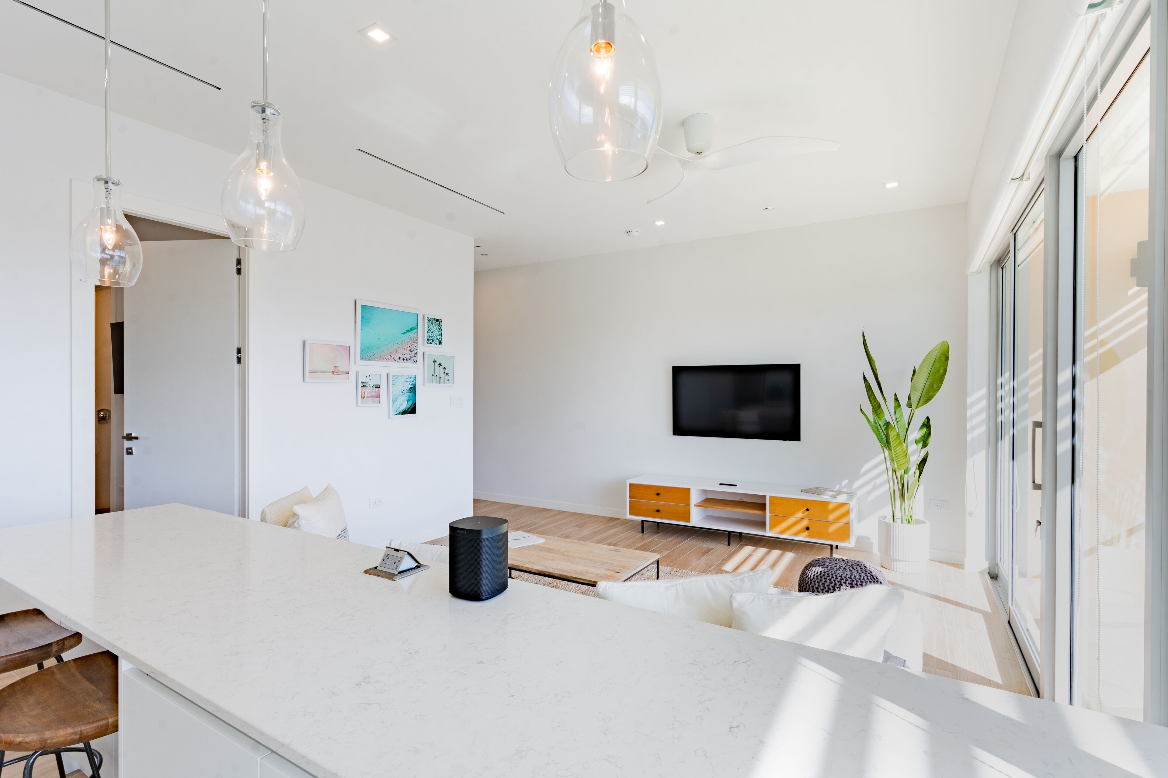 A modern living room with a white sofa, a coffee table, and a TV mounted on the wall. There is a plant on a side table and a large window providing natural light.