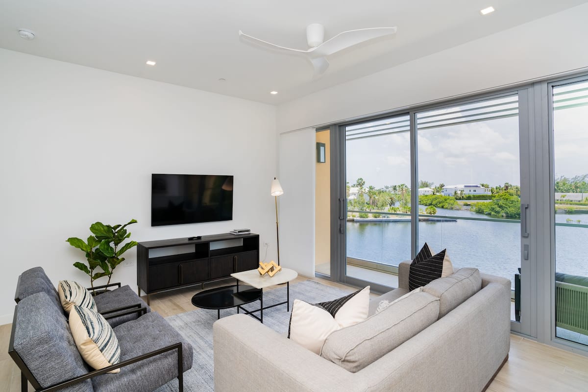 A modern living room with a large TV, comfortable seating, a small table, and a view of a body of water through large windows.