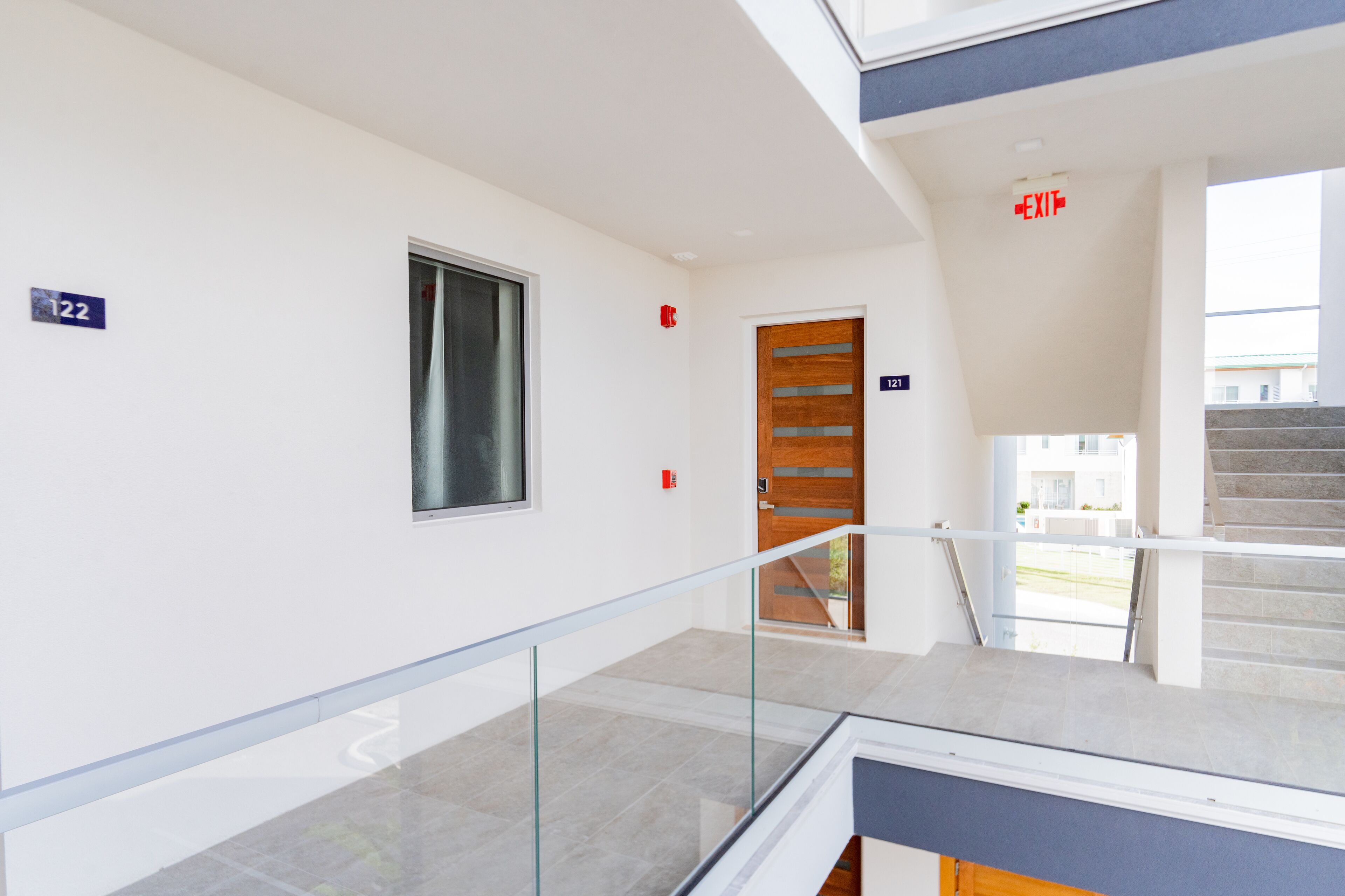 A modern hotel corridor with a glass railing, a wooden door, and a staircase leading to an outdoor area.