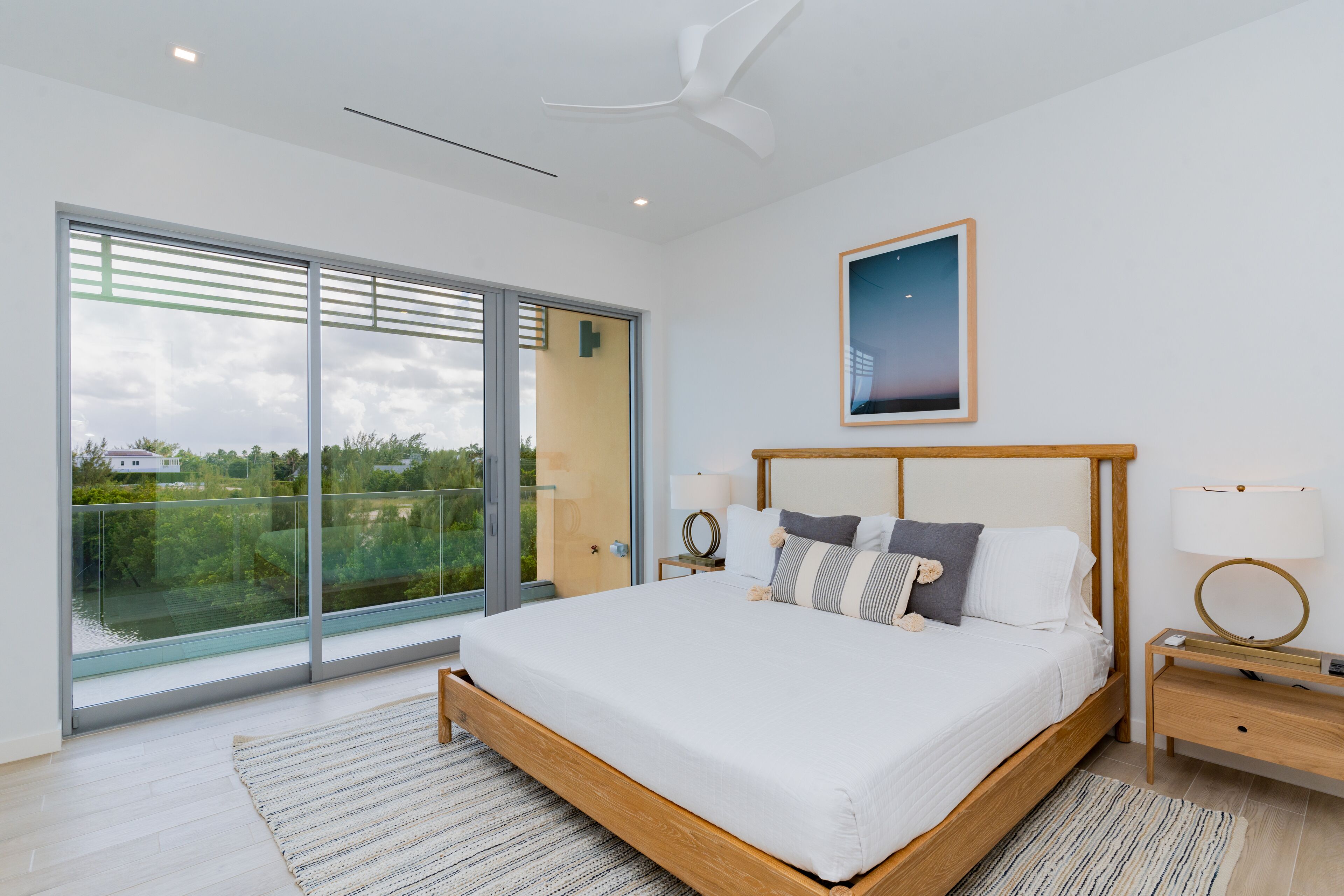 A modern bedroom with a large bed, a ceiling fan, a nightstand, and a view through a sliding glass door.
