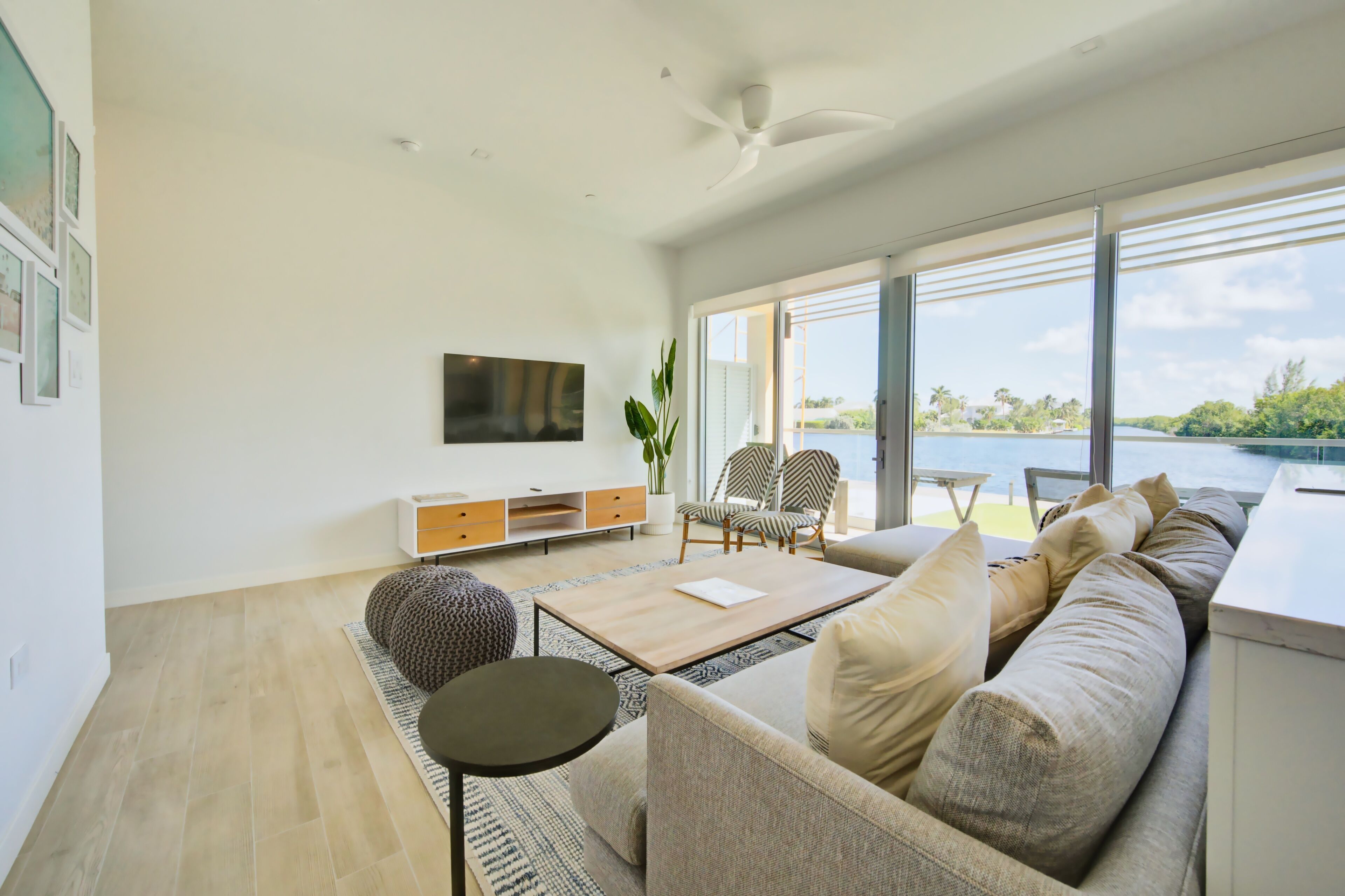 A modern living room with a sofa, coffee table, and chairs, large windows with a view of the water, and a flat-screen TV mounted on the wall.