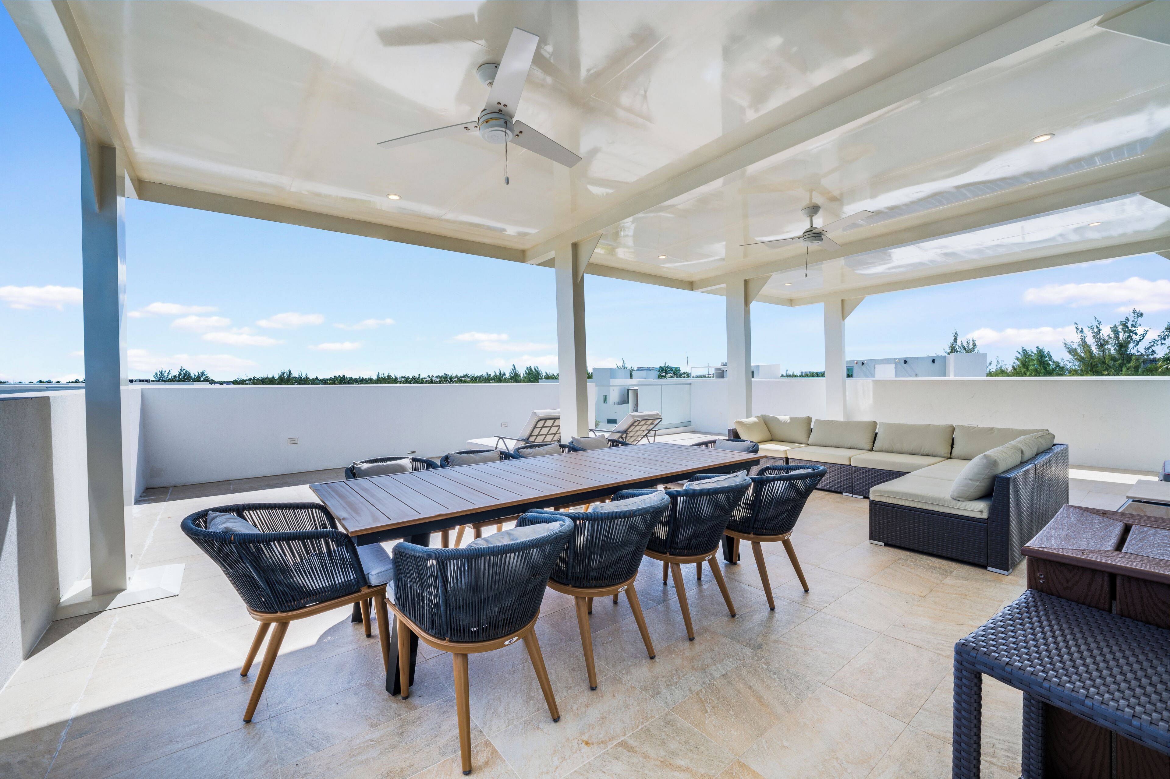 Un espacio exterior techado con zona de comedor, una mesa larga y sillas, un sofá y un ventilador de techo.