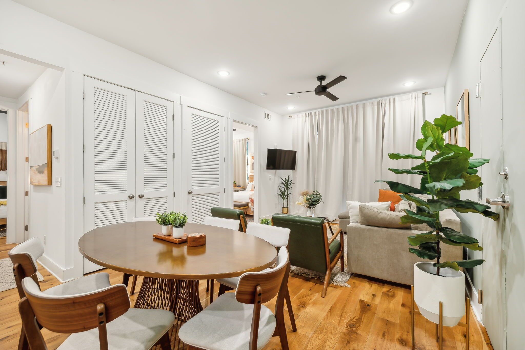 A modern living room with a round wooden table, green and white chairs, a sofa, and a large potted plant.