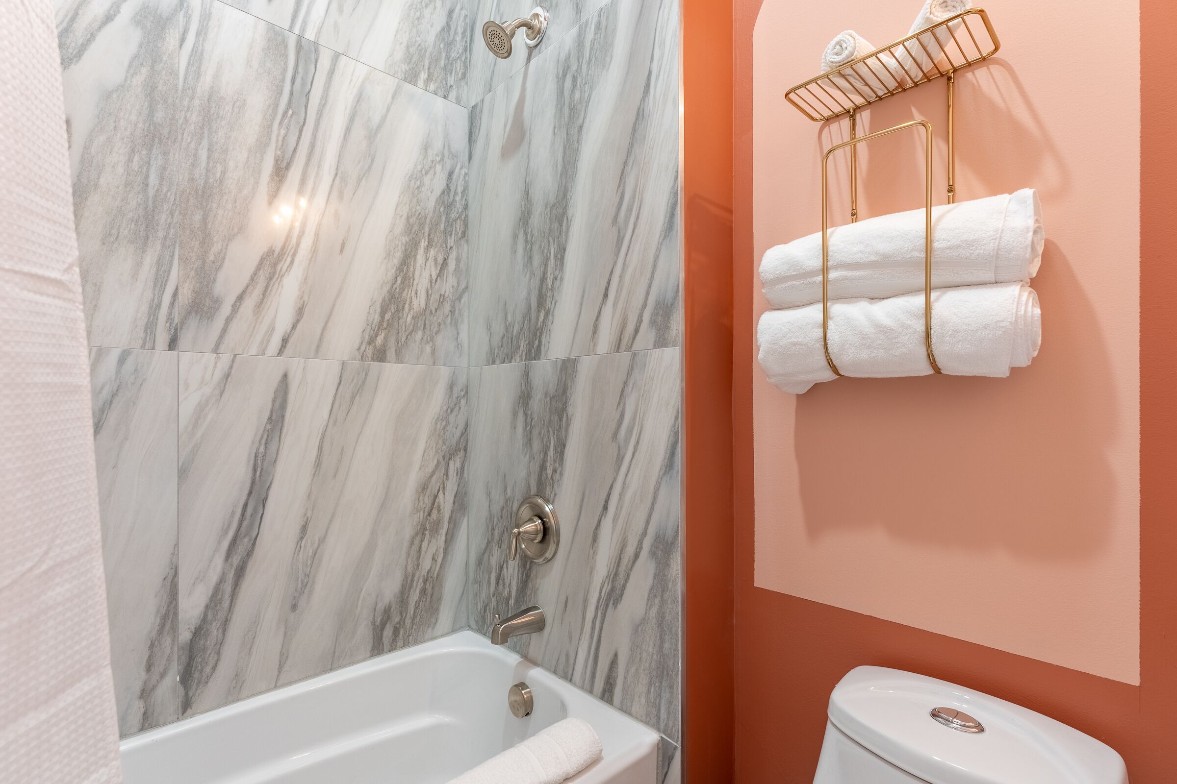 A bathroom with a marble shower wall, a white bathtub, and a toilet. There are towels hanging on a gold rack.