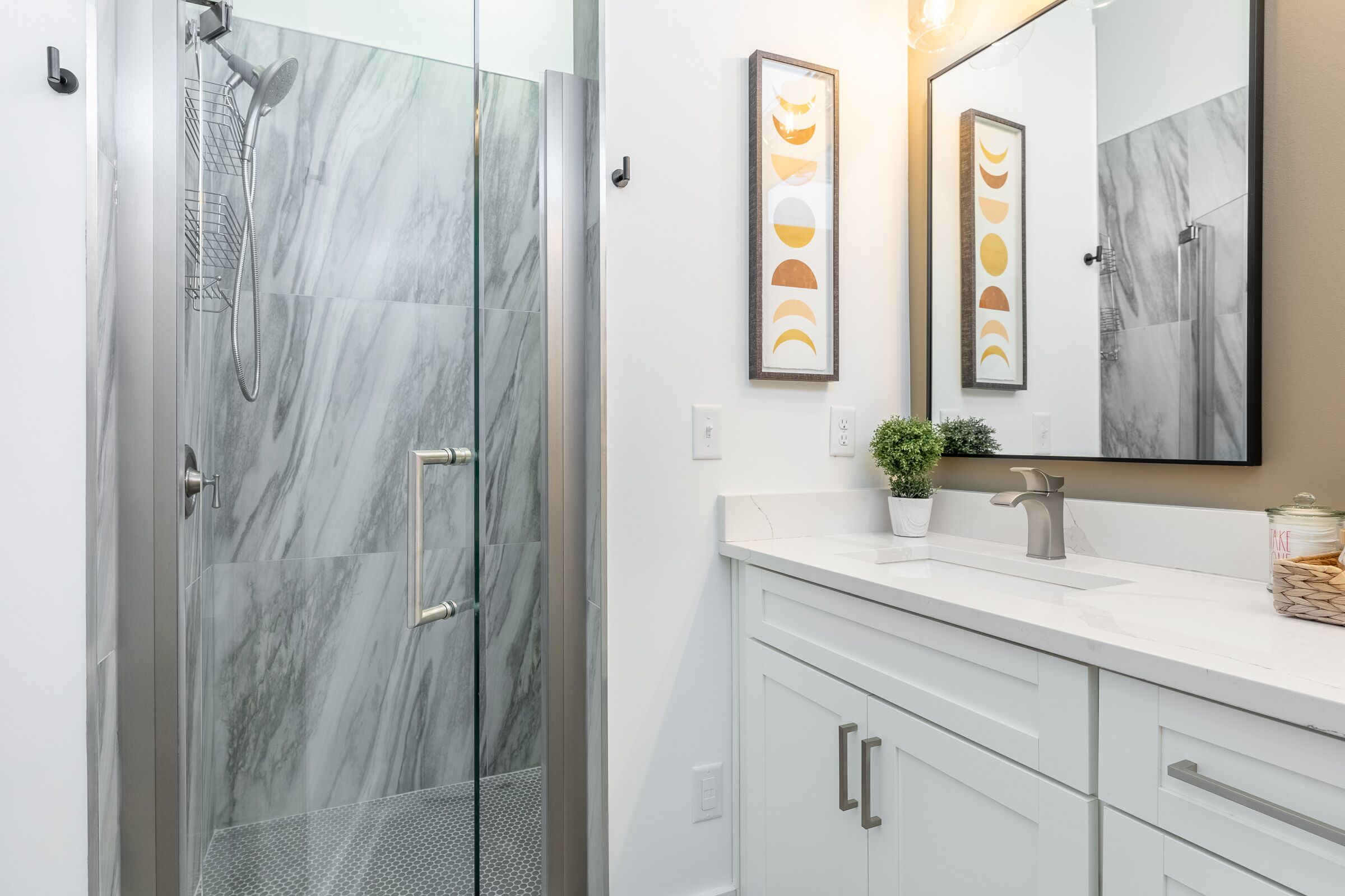 A modern bathroom with a glass-enclosed shower, white marble tiles, a white vanity with a sink, and a framed artwork above the mirror.