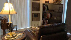 Ein Wohnzimmer mit Ledersofa, einem Tisch mit Lampe, einem Bücherregal und einem Fenster mit Jalousien.