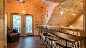 Interior acogedor de una cabaña de madera, con una zona de bar y un área de estar con cojines, y un cartel que dice 'Believe'.