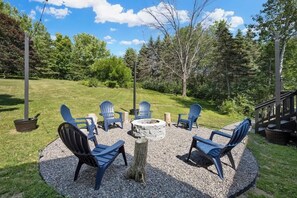 A circular seating area with Adirondack chairs around a stone fire pit, set in a gravel circle with a grassy lawn in the background.