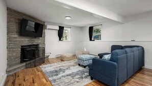 A modern living room with a stone fireplace, a large flat-screen TV mounted on the wall, a blue sofa, and a patterned rug on a wooden floor. - Finger Lakes ~ Firepit, Spacious, Wine Country (Skaneateles)