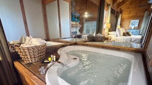 A large, freestanding bathtub in a wooden cabin-style bathroom with a stone wall and a large mirror reflecting the room. - Couples Retreat | In-Bedroom Jacuzzi, Fireplace & Private Deck · Near Helen GA (Sautee Nacoochee)