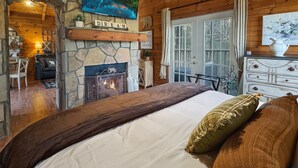 A cozy bedroom with a stone fireplace, a large bed, and a scenic painting above the fireplace. - Couples Retreat | In-Bedroom Jacuzzi, Fireplace & Private Deck · Near Helen GA (Sautee Nacoochee)