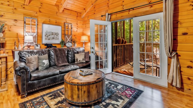 A cozy cabin interior with a leather sofa, a round wooden coffee table, and a large sliding glass door leading to an outdoor deck. - Couples Retreat | In-Bedroom Jacuzzi, Fireplace & Private Deck · Near Helen GA (Sautee Nacoochee)