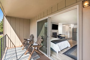 Uma varanda com cadeiras de madeira e um tapete listrado, uma porta de vidro deslizante que dá para um quarto com uma cama, televisão e frigorífico.