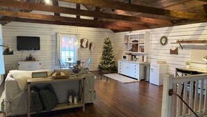 A cozy living room with a wooden ceiling, a TV mounted on the wall, a Christmas tree, and a dining area with a white cabinet and a clock.