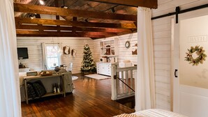 A cozy room with a wooden ceiling, a bed with a striped blanket, a small table with a basket, a Christmas tree, and a kitchen area with a microwave.