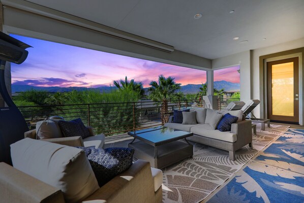 Un área de estar exterior moderna con sofá, sillas y una mesa de centro, con vistas a un paisaje pintoresco con montañas y un atardecer.