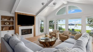 A spacious living room with a large flat-screen TV, a fireplace, and a view of a lake and mountains. - VRBO 2025 Rental of the Year | Folly Riverfront Retreat w/ Dock & Pool (Folly Beach)