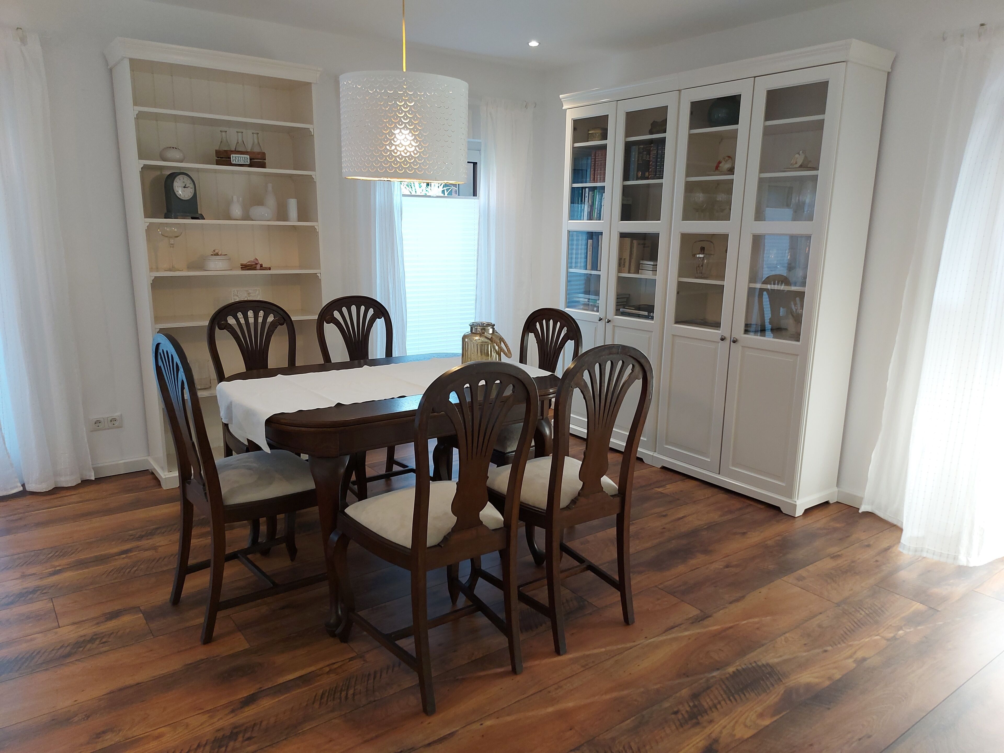 A dining room with a wooden table and chairs, white walls, and a cabinet with glass doors.