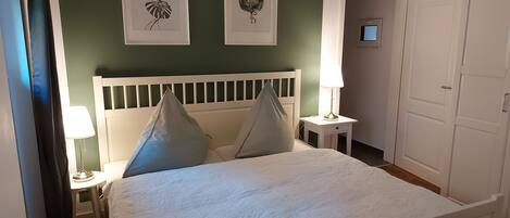 A bedroom with a bed, two pillows, a nightstand with a lamp, and two framed botanical prints on the wall.