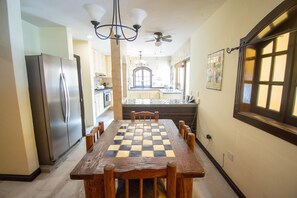 A dining area with a checkered table, wooden chairs, a refrigerator, and a ceiling fan.