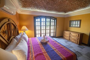 A hotel room with a bed, a wooden dresser, a window with bars, and a ceiling with exposed brick.