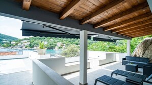 A covered outdoor area with a wooden ceiling, a seating area with sofas and tables, and a view of a coastal landscape with buildings and greenery. - Hidden Gem Apartment - Two Bedroom Apartment with Terrace and Sea View (Molunat)