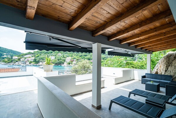 A covered outdoor area with a wooden ceiling, a seating area with sofas and tables, and a view of a coastal landscape with buildings and greenery.