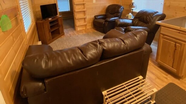 A cozy cabin interior with a brown leather sofa, a ceiling fan, wooden walls, and a small TV. - Log Cabin by ND Badlands (Grassy Butte)