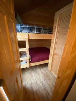 A small, wooden bunk bed room with a single bed and a lower bunk. There is a wooden door on the right and a small shelf under the bed.