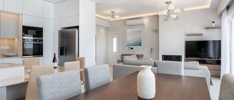 Et moderne køkken med indbyggede apparater, et spiseområde med et træbord og grå polstrede stole, samt en opholdsstue med sofa og fjernsyn.