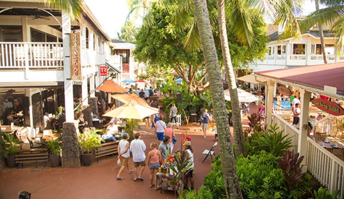 An outdoor market area with people walking and sitting under umbrellas, surrounded by palm trees and buildings with balconies.