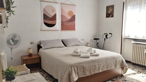 A neatly arranged bedroom with a bed, bedside tables, a standing fan, a potted plant, and artwork on the wall. - Charming Perledo Retreat with Lake View, Balcony & Parking (Perledo)