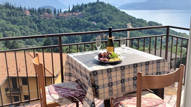 A balcony with a table set for two, overlooking a scenic view of mountains and a lake. - Charming Perledo Retreat with Lake View, Balcony & Parking (Perledo)