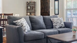 A gray sofa with patterned pillows, a wooden coffee table, and a bookshelf in a room with a wooden door and window blinds. - La Casa Roja - Pet-Friendly - 3BR Home - Walk to Castle & Downtown Waco (Waco)