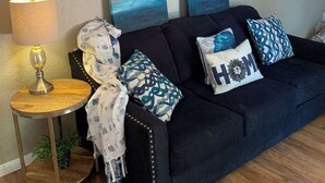 Un salon avec un canapé bleu foncé, deux œuvres d’art bleues accrochées au mur, une petite table ronde en bois avec une lampe, et des coussins décoratifs à motifs.